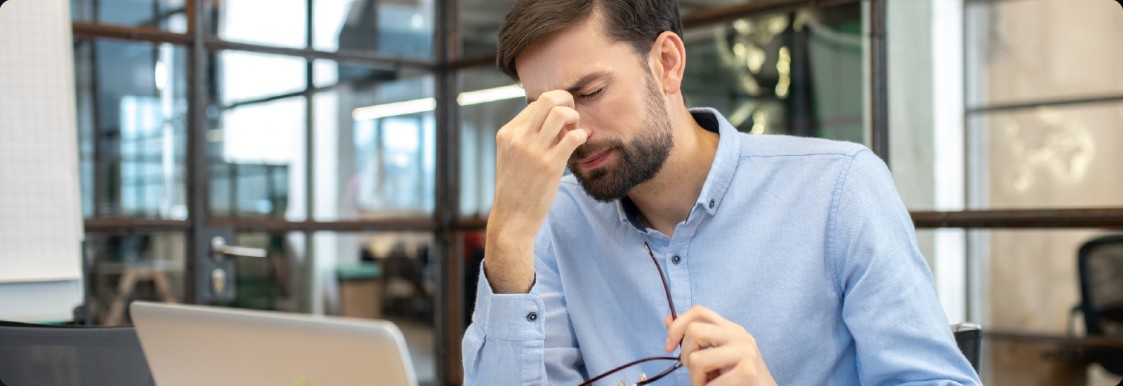 A man at a laptop, holding his glasses and rubbing his eyes